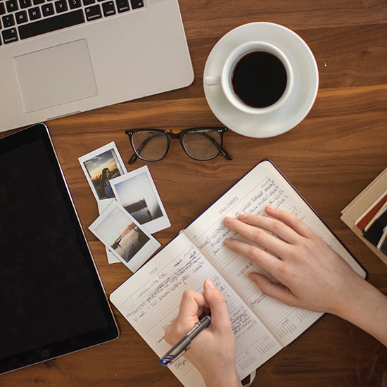 birdseye view of someone writing in a journal at a desk with a pair of glasses and a cup of coffee